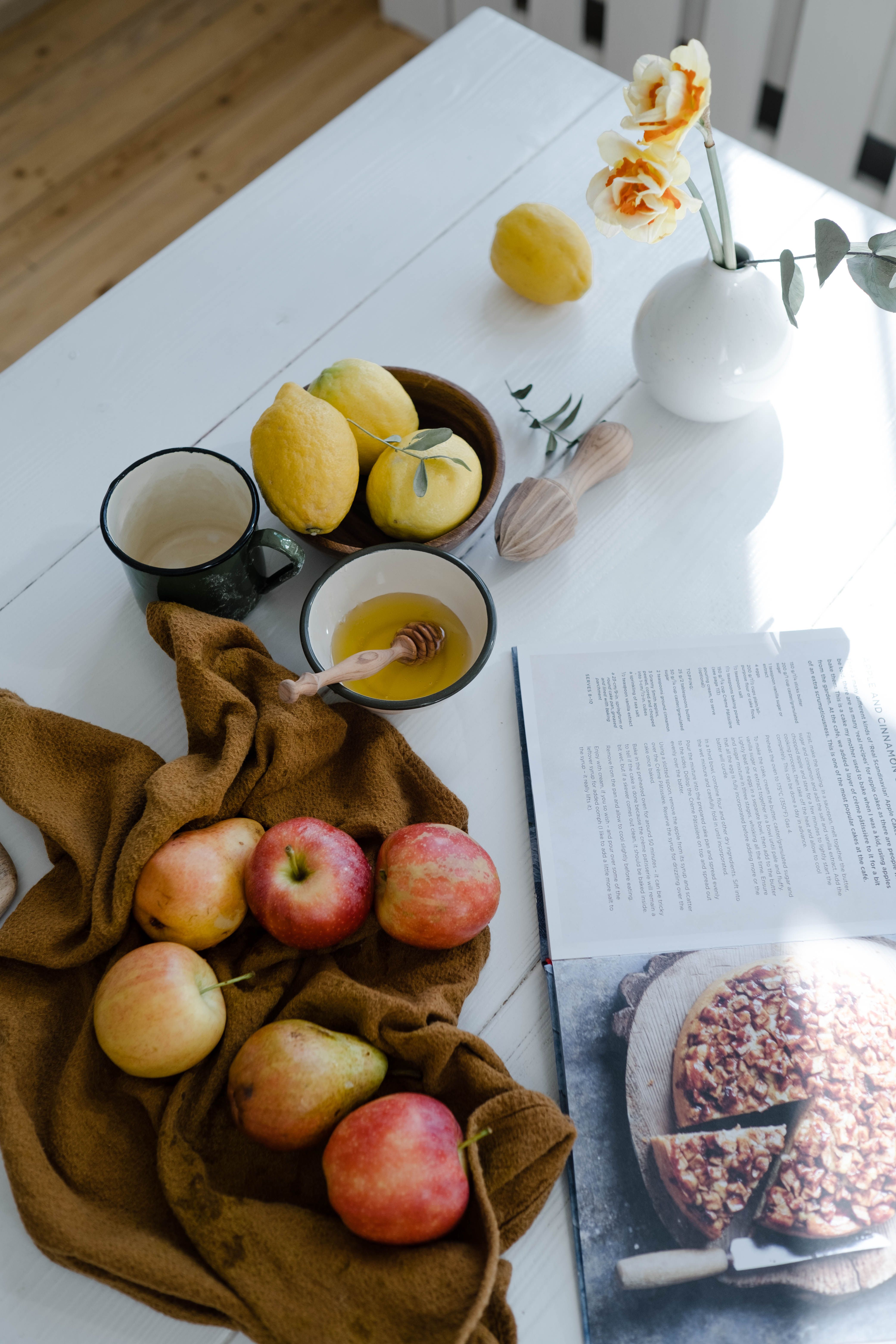 Open cookbook beside apples, pears, lemons and honey on a kitchen table, illustrating the contrast between British and Mediterranean food traditions.